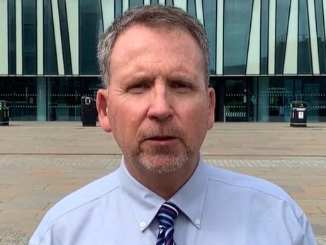 Head and shoulders image of a man in a blue shirt with a glass building behind him