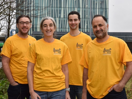 Four people in yellow t-shirts standing on grassland with the Sir Duncan Rice Library in the background