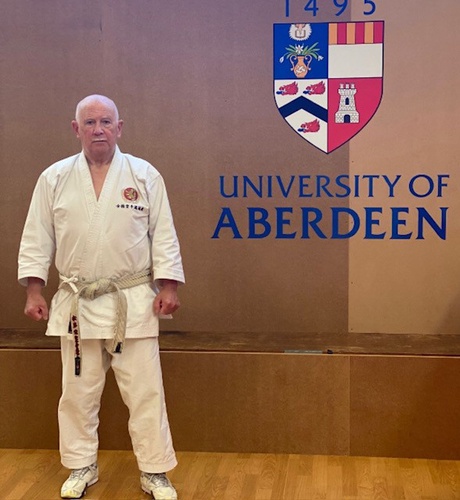 Ronnie Watt stands in his Karate kit by the University of Aberdeen logo