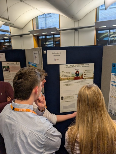 A man and woman stood looking at student Joe's research poster titled &ldquo;I Will Walk 500 Metres, and a Pheasant Will Walk 500 More&rdquo;