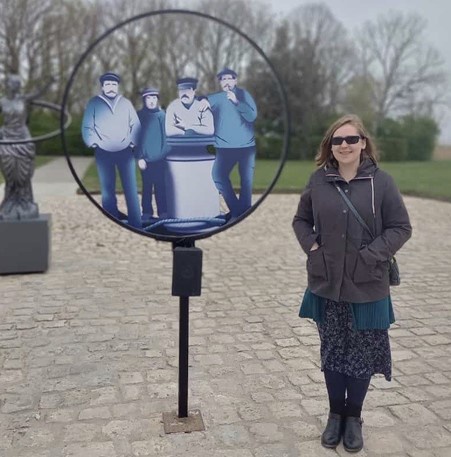 A pole sticking out from a paved road with a hoop at the top and four sailors depicted within the hoop. A woman in a dark coat and sunglasses is stood next to the pole.