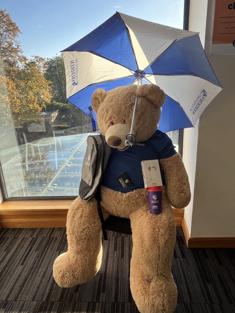 A big stuffed teddy holding the prizes: earphones, an umbrella, coffee cup, power bank and laptop bag