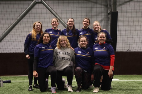 9 people in sports attire smiling on an indoor football pitch.