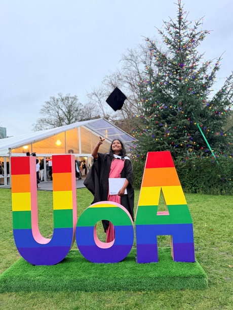 Angara Gowthami standing behind UoA letters, throwing their graduation cap in the air