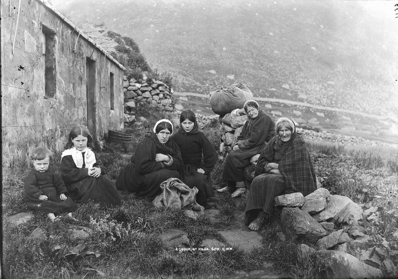 Black and white photograph of women and children outside a homestead in the hills. The women are wearing dresses and tartan, but no shoes, and sit upon a stone wall