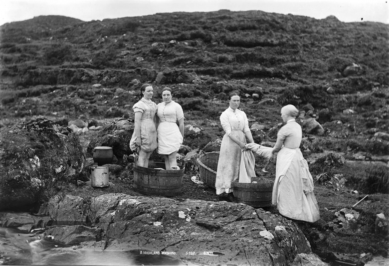 Black and white photograph showing four women in long dresses in the hilld. Two are standing in a bucket holding their skirts to their knees while the other two are ringing out a piece of cloth. There are rock pools and rocky hills behind them.