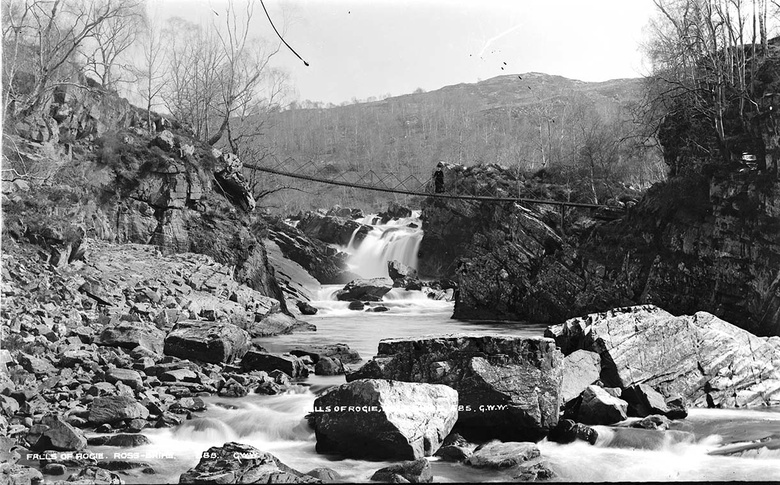 Black and white photograph of a meandering river with a waterfall in the background. The river is broken up with large rocks and a rope bridge crosses above it