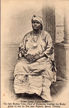 Image of Madam Yoko sitting on a chair wearing a full length clothing and head scarf, with medal around her neck. Her hands are clasped in her lap.