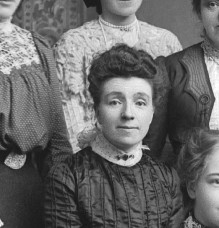 Close up of one of the women from the group photograph. She has dark hair in a bun and is wearing a black dress with a white lace collar