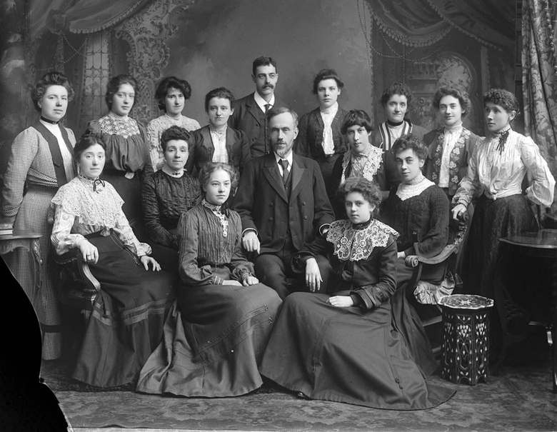 Black and white photograph of a group of women, and two men, stting in three rwos. The figures wear Victorian clothing and are against a backdrop of curtains