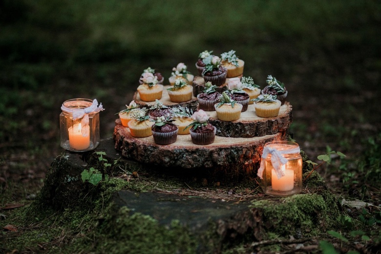 Cupcakes piled on a wooden slab in a forest with candles surrounding them