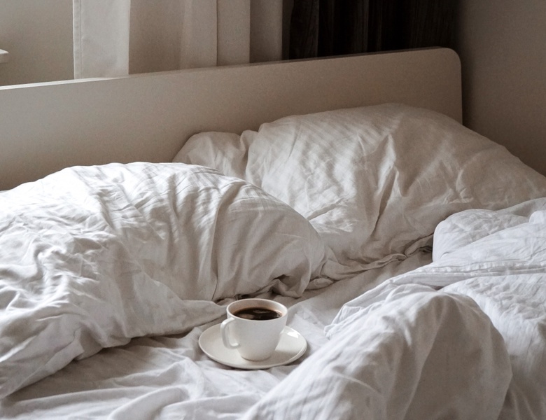 A cup of coffee on a saucer sits in the middle of an unmade bed with crisp white sheets